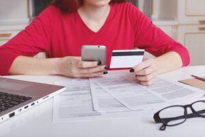 cropped shot of unrecognizable female model in red sweater sits at working table surrounded with papers and laptop computer, uses cell phone for paying bill online with credit card. ecommerce concept